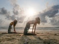 Two women practice camel yoga asana at seashore in sunlight