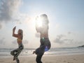 Two women practice eagle yoga asana at seaside in sunlight