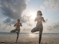 Two women practice tree yoga asana at seaside in sun light