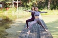 Two women practicing yoga in the morning on a wooden bridge in the park perform virabhadrasana exercise, warrior pose Royalty Free Stock Photo