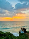 Two women sit side by side near a rocky cliff, overlooking an awe-inspiring sunset on the seashore