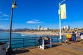 Two women sitting on a bench on a wooden pier with vast blue ocean water, the cityscapes along the beach, mountain ranges