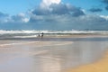 Two women tourists- one with arms outstretched enjoy standing out on wet beach under stormy sky with turbulent ocean in background