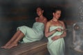 Two women in a traditional steam sauna banya pours water on hot stones on the stove