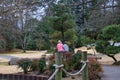 Two women walking in a Japanese garden surrounded by lush green trees and plants with yellow winter grass