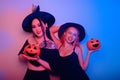 Two women witches hold pumpkins in their hands and dance. Women in neon lights are celebrating a Halloween party. Models smile and