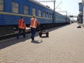 Two workers of the iron orog are pushing a cart around the platform. Workers in overalls are walking along a passenger carriage of