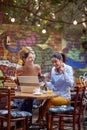 Two young adult women sitting in outdoor cafe, smiling, laughing, having online communication with someone