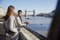 Two young adult women standing at the Thames riverside talking, Tower Bridge in the background