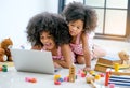 Two young African girls play with notebook computer among toys, doll and book in front of glass window