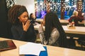 Two young african women gossip at table. On of them looks wondered. Another model keep finger at mouth. They have laptop