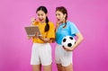 Two Young Asian woman over isolated pink background holding a soccer ball and laptop, whatching football match, sport concept