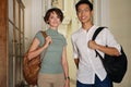 Two young attractive international students with backpacks happily looking in camera while standing in corridor of