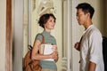 Two young attractive international students with backpacks happily talking while standing in corridor of university