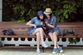 Two young beautiful girls in jeans dresses and hats sit on a bench in the park on a background of green plant walls, and Royalty Free Stock Photo