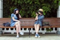 Two young beautiful girls in jeans dresses and hats sit on a bench in the park on a background of green plant walls, and Royalty Free Stock Photo