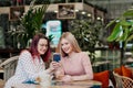 Two young beautiful girls sit in a green cafe at a table. Drink tea with croissants, chatting, laughing and taking Royalty Free Stock Photo