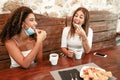 Two young beautiful mixed-race women sitting outdoors on rustic wooden table wearing lowered protective mask eating some pastries Royalty Free Stock Photo