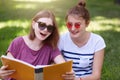 Two young beautiful smiling women reading somethig interesting, sitting on grass at summer green park, prepare for exam toogether Royalty Free Stock Photo