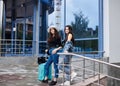 Two young brunette girls, wearing casual jeans attire, standing with mint luggage in front of blue modern glass building