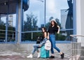 Two young brunette girls, wearing casual jeans attire, standing with mint luggage in front of blue modern glass building