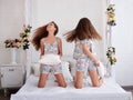 Two young brunette women girls, wearing cotton pajamas, sitting on white bed in light room apartment, smiling, having pillow fight