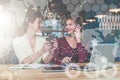 Two young businesswomen are sitting at table.First girl is holding smartphone,second is talking on cell phone