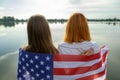 Two young friends women with USA national flag on their shoulders standing together outdoors on lake shore. Patriotic girls Royalty Free Stock Photo