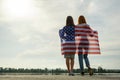 Two young friends women with USA national flag on their shoulders standing together outdoors on lake shore. Patriotic girls Royalty Free Stock Photo