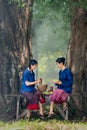 Two young girls prepare food using motar and other flavoring look like to make papaya salad and they sit under big tree with day