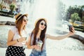 Two young girls in sunglasses and hats near city fountain. Best friends drink cocktails doing self photo and laughing in city stre