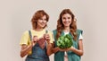 Two attractive young girls, twin sisters in casual wear holding fresh green salad and piece of meat isolated over light