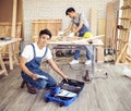 Two handsome Asian male repairmen or carpenters working together in wood workshop, one man sitting smiling and looking at camera