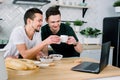 Two young handsome men having breakfast and drinking coffee while using laptop in the kitchen at the morning. Smiling Royalty Free Stock Photo