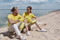 Two young happy eco activists or volunteers wearing uniform and rubber gloves sitting on the beach and discussing Royalty Free Stock Photo