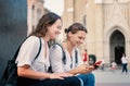 Two young happy pretty female students use a smartphone while sitting on the square in the city of Europe