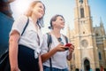 Two young happy pretty female students use a smartphone while sitting on the square in the city of Europe