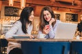 Two young happy women are sitting in cafe at table in front of laptop, using smartphone and laughing. Royalty Free Stock Photo