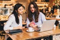 Two young happy women are sitting in cafe at table in front of laptop, using smartphone and laughing. Royalty Free Stock Photo