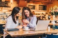 Two young happy women are sitting in cafe at table in front of laptop, using smartphone and laughing. Royalty Free Stock Photo