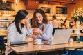 Two young happy women are sitting in cafe at table in front of laptop, using smartphone and laughing. Royalty Free Stock Photo