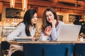 Two young happy women are sitting in cafe at table in front of laptop, using smartphone and laughing. Royalty Free Stock Photo