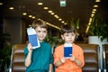 Two young kids sitting on brown chairs, holding passports and tickets in waiting hall in airport. Travel and holidays with Royalty Free Stock Photo