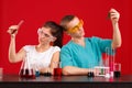Two lab technicians, a girl and a guy, are holding bottles with a multicolored liquid looking at it. On a red background