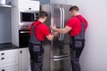 Two Young Male Movers Placing Steel Refrigerator In Kitchen