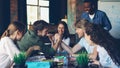 Two young men are competing in arm wrestling during break at work while their coworkers are cheering, laughing and