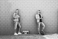 Two young man looking at mobile phone lean against brick wall - Couple of cool guys listening music with vintage boombox stereo Royalty Free Stock Photo
