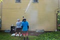 Two young men standing by the side of a yellow house. One is power washing the siding with a long nozzle