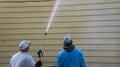 Two young men standing by the side of a yellow house. One is power washing the siding with a long nozzle