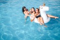 Two young models swimming on unicorn shaoed float. They smiles and laugh. Woman in front holds cocktail in hands. Thirl